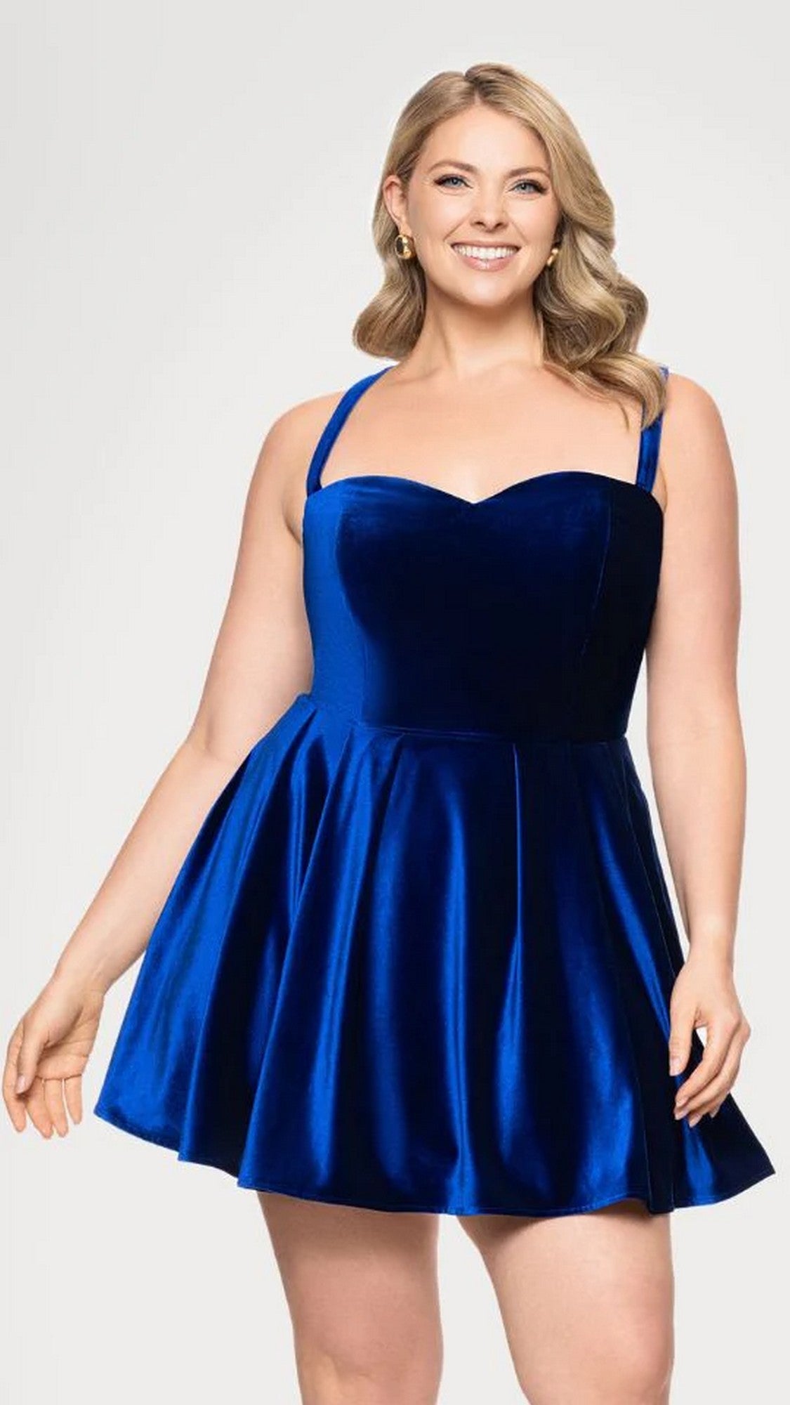 A woman wears the Short Plus-Size Prom Dress Blondie Nites 4469BNW in sapphire, featuring a sleeveless fit-and-flare velvet design, as she stands smiling against a plain light background.